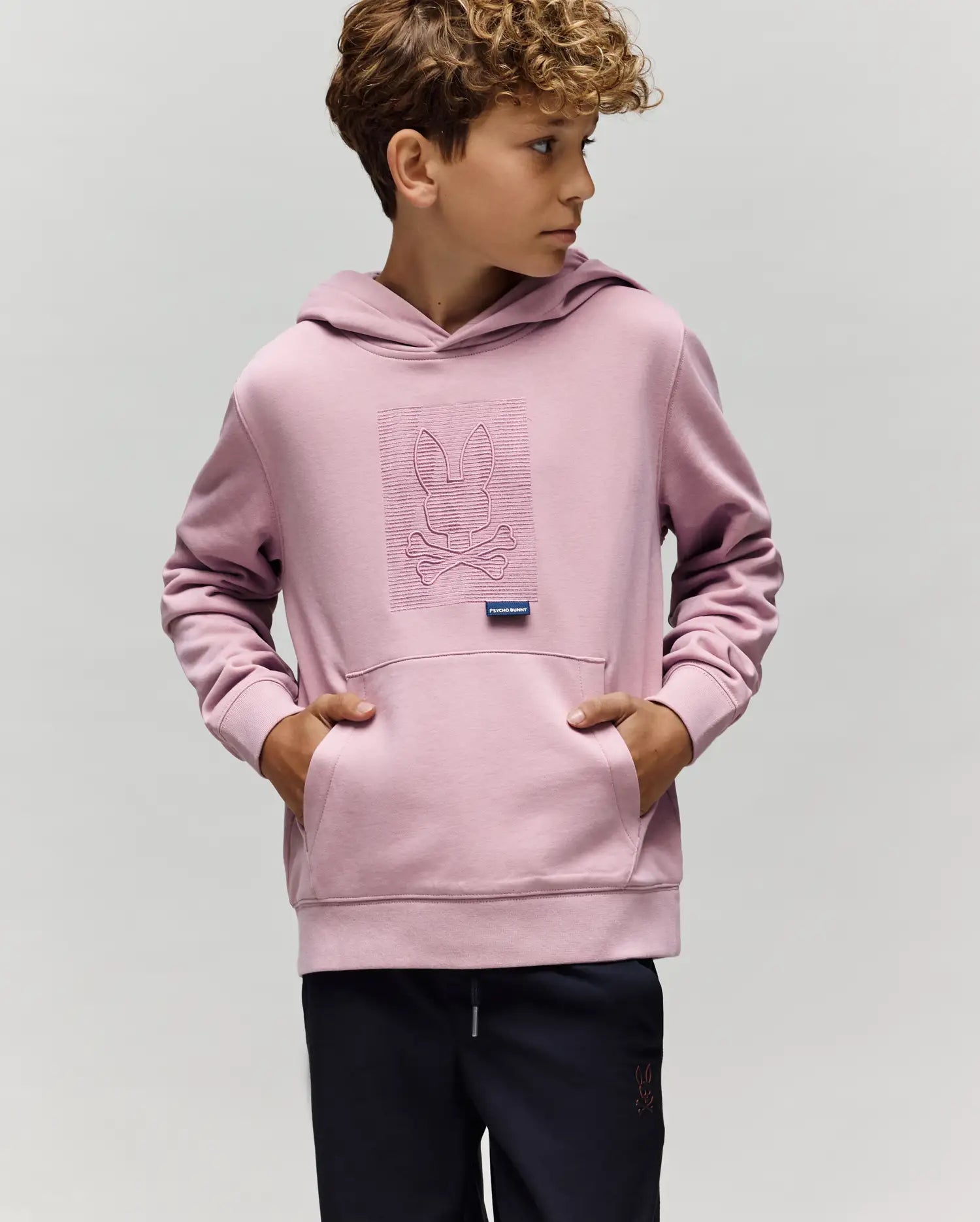 A young boy with curly hair wears the Psycho Bunny KIDS GRANT HOODIE - KT0500020 in light purple, featuring an embroidered bunny and crossbones. He stands with hands in the front pocket, looking right against a plain background.