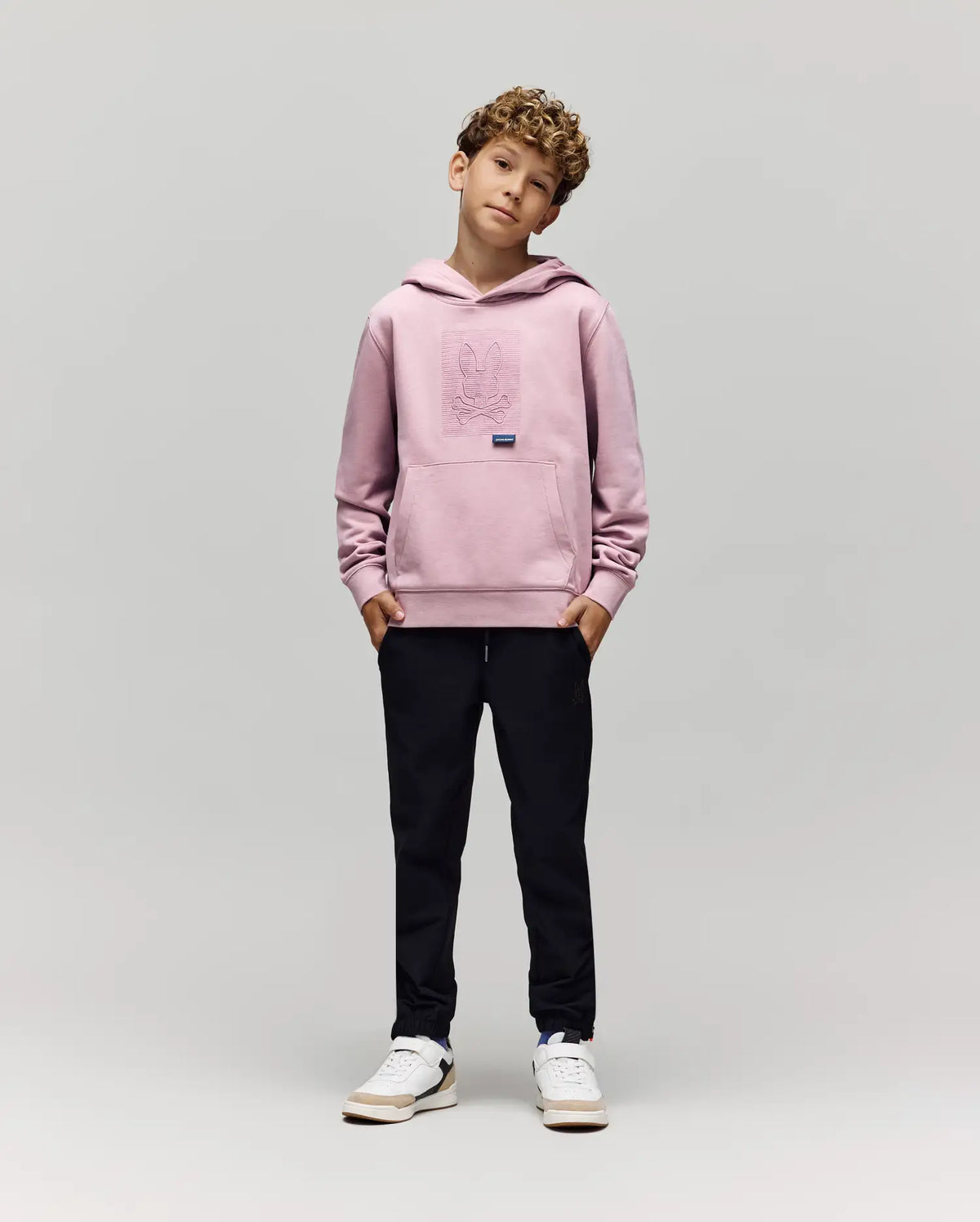 A young boy with curly hair stands with hands in pockets, wearing the Psycho Bunny KIDS GRANT HOODIE - KT0500020 in light pink, paired with black pants and white sneakers against a plain light gray background.