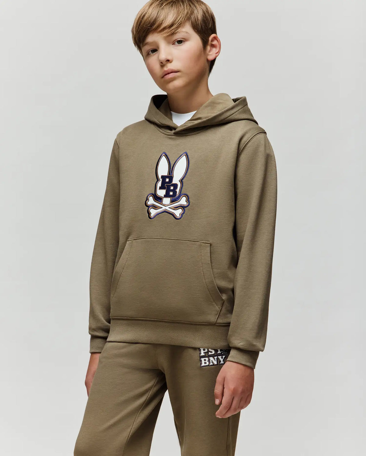 A boy in a Psycho Bunny KIDS WADE CORD PATCH HOODIE - KT0500031, an olive green cotton blend athletic hoodie with an embroidered bunny patch, and matching sweatpants stands against a plain light gray background.