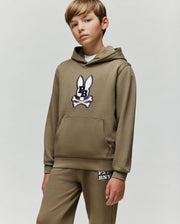 A boy in a Psycho Bunny KIDS WADE CORD PATCH HOODIE - KT0500031, an olive green cotton blend athletic hoodie with an embroidered bunny patch, and matching sweatpants stands against a plain light gray background.