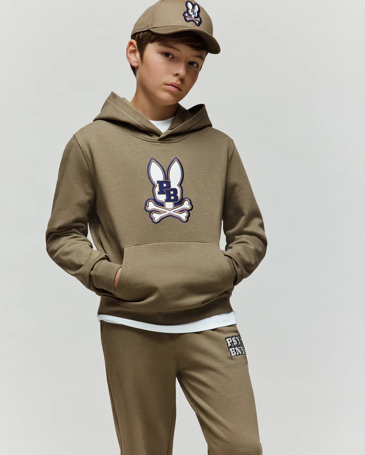 A boy wears the Psycho Bunny KIDS WADE CORD PATCH HOODIE (KT0500031) and matching joggers, featuring an embroidered bunny patch, khaki cap, and white shirt, standing with one hand in his pocket against a plain light background.