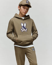 A boy wears the Psycho Bunny KIDS WADE CORD PATCH HOODIE (KT0500031) and matching joggers, featuring an embroidered bunny patch, khaki cap, and white shirt, standing with one hand in his pocket against a plain light background.