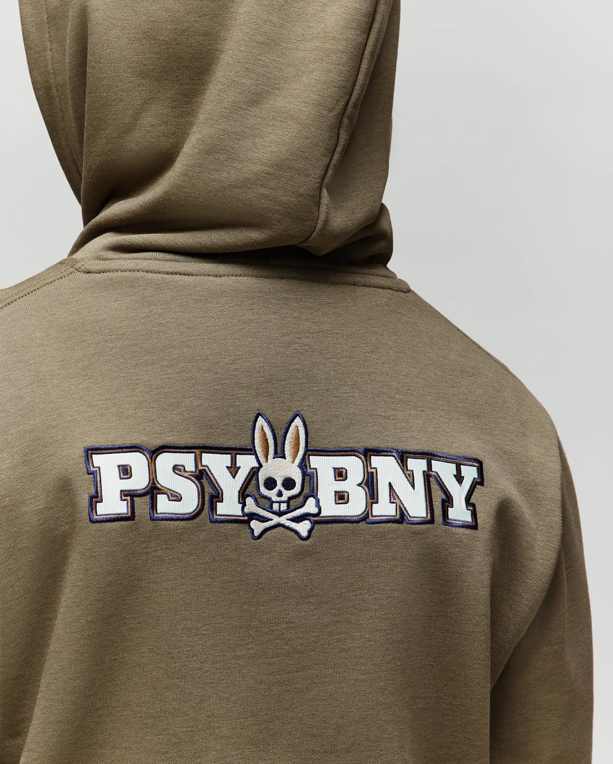 A person wearing the Psycho Bunny KIDS WADE CORD PATCH HOODIE (KT0500031) in olive green, featuring "PSYBUNY," a skull with bunny ears and crossed bones logo on the back, and an embroidered bunny patch.