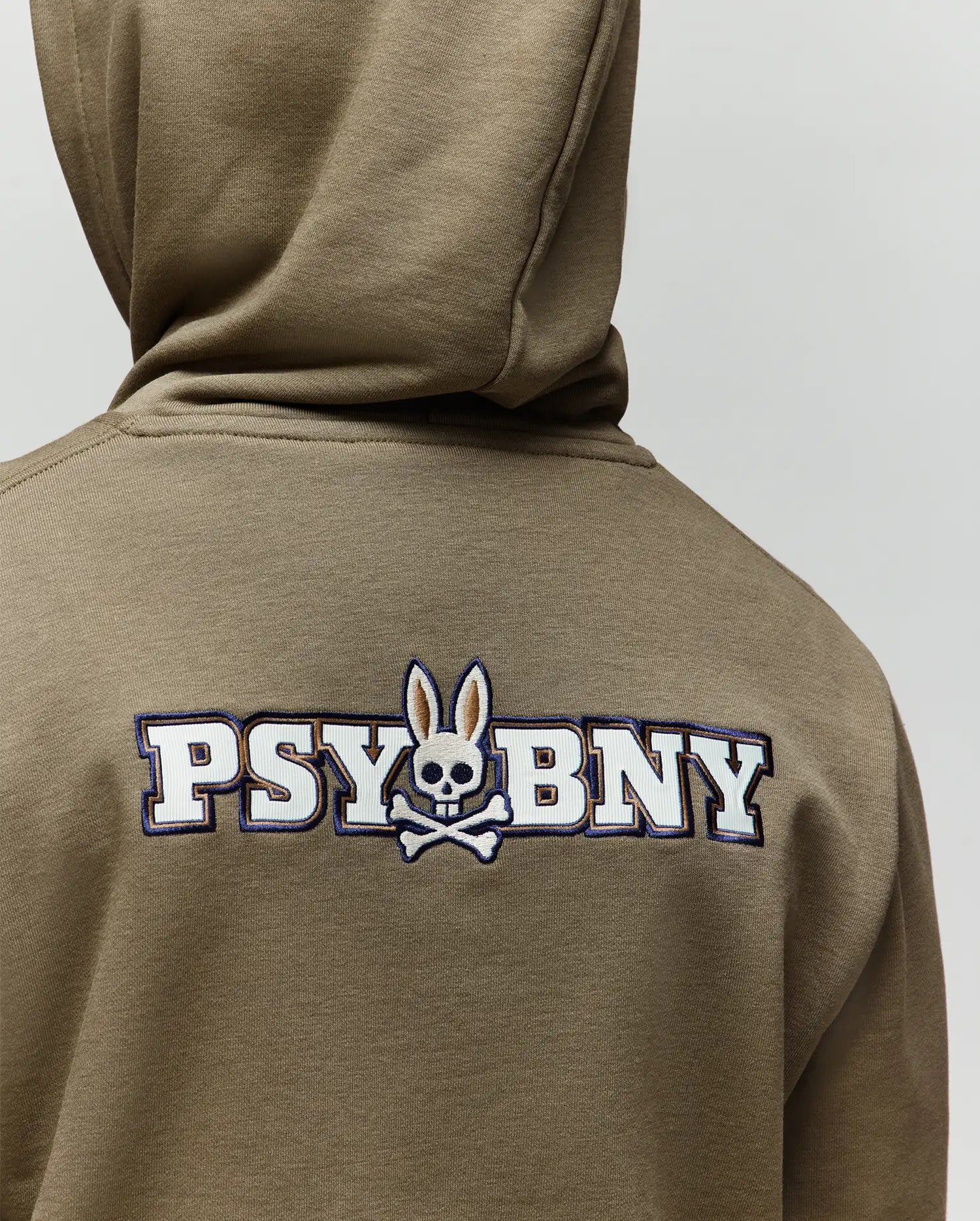 A person wearing the Psycho Bunny KIDS WADE CORD PATCH HOODIE (KT0500031) in olive green, featuring "PSYBUNY," a skull with bunny ears and crossed bones logo on the back, and an embroidered bunny patch.