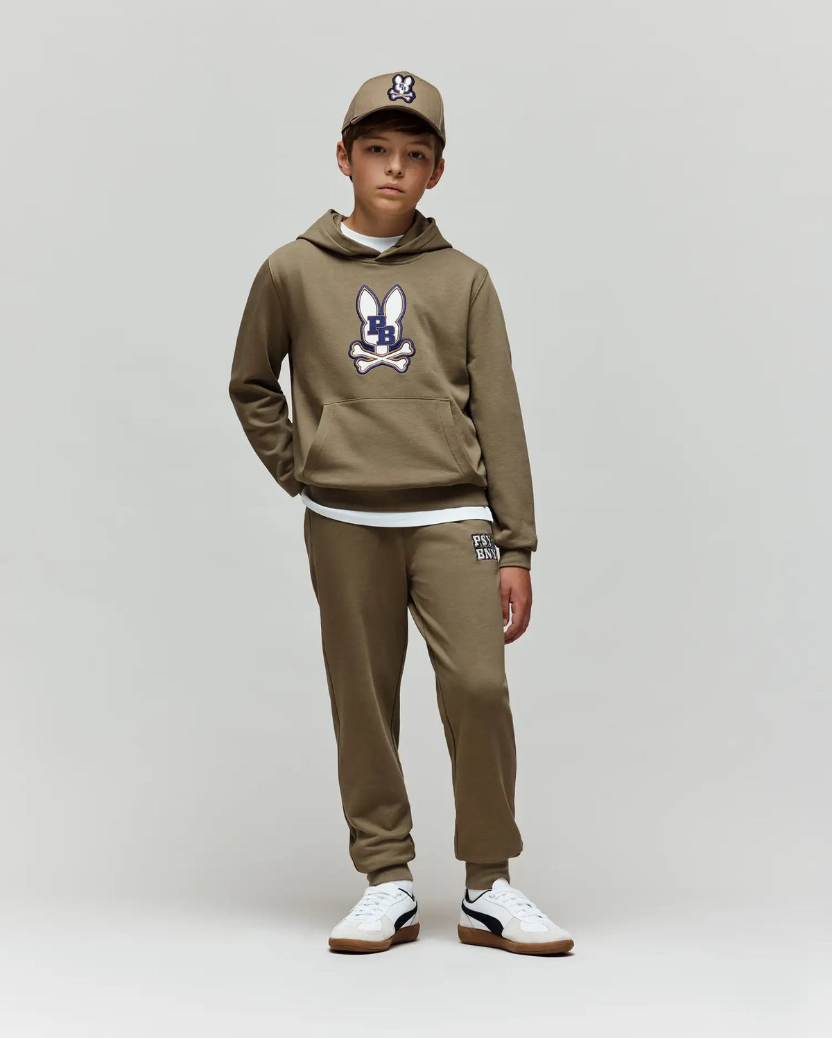 A boy stands against a plain background wearing the Psycho Bunny KIDS WADE CORD PATCH HOODIE (KT0500031) in olive green, matched with sweatpants, a cap, white undershirt, and white sneakers with black accents.