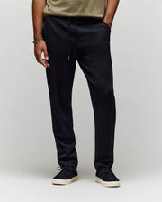A person wearing Psycho Bunny MENS ALEXIS PINTUCK JOGGER - MB0800006 in black, paired with a beige shirt and black sneakers with white soles, stands against a plain light background. Only the lower half is visible.