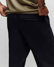 A person wears Psycho Bunny's black MENS ALEXIS PINTUCK JOGGER (MB0800006) with "PSYCHO BUNNY" embroidered above the back pocket, an olive green shirt, and their left hand on their hip. The joggers feature subtle pintuck detailing.