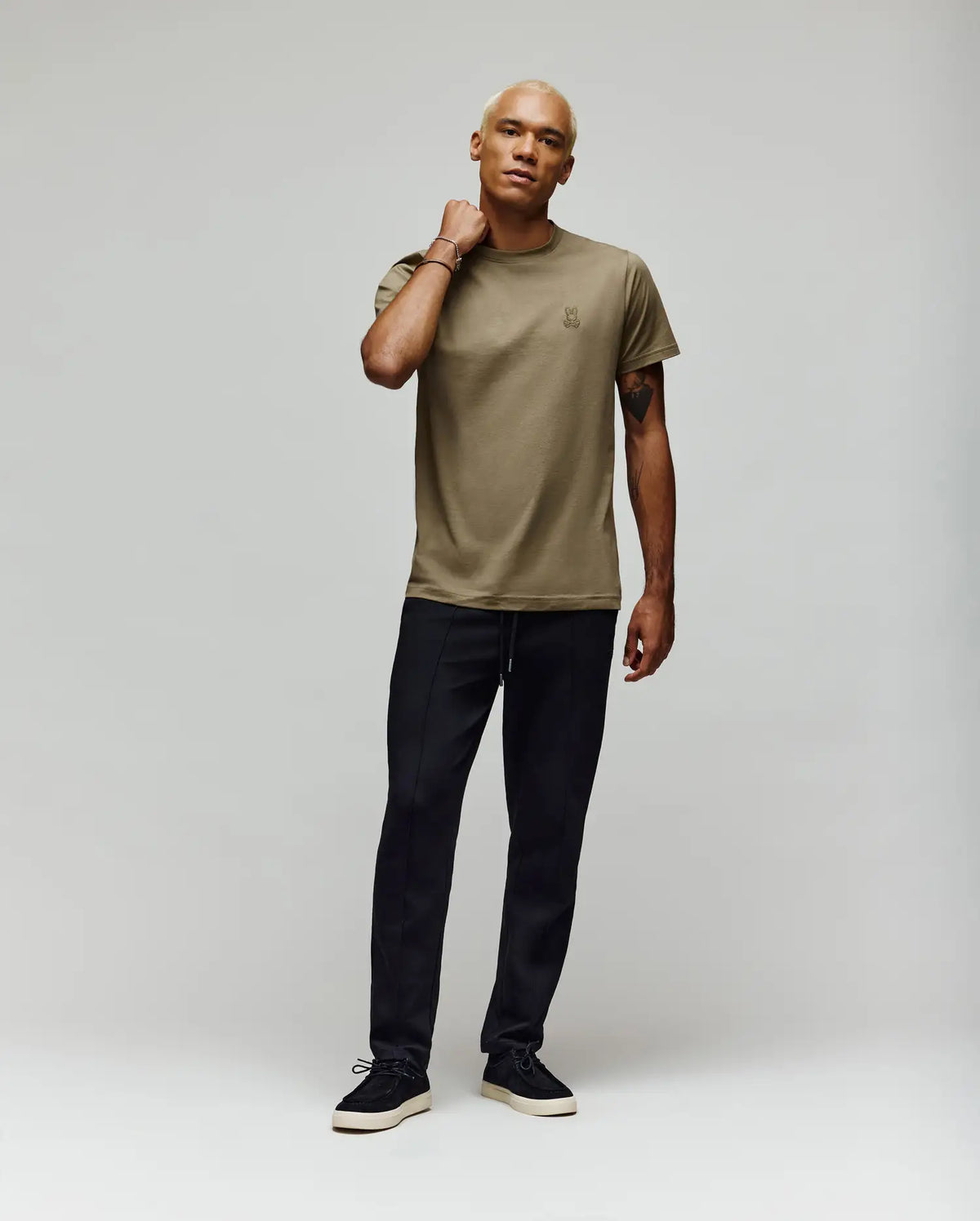 A man with short blonde hair stands against a plain light background, wearing an olive green t-shirt and Psycho Bunny MENS ALEXIS PINTUCK JOGGER - MB0800006 in black, looking relaxed with one hand on his neck and wearing black sneakers.