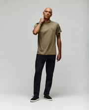 A man with short blonde hair stands against a plain light background, wearing an olive green t-shirt and Psycho Bunny MENS ALEXIS PINTUCK JOGGER - MB0800006 in black, looking relaxed with one hand on his neck and wearing black sneakers.