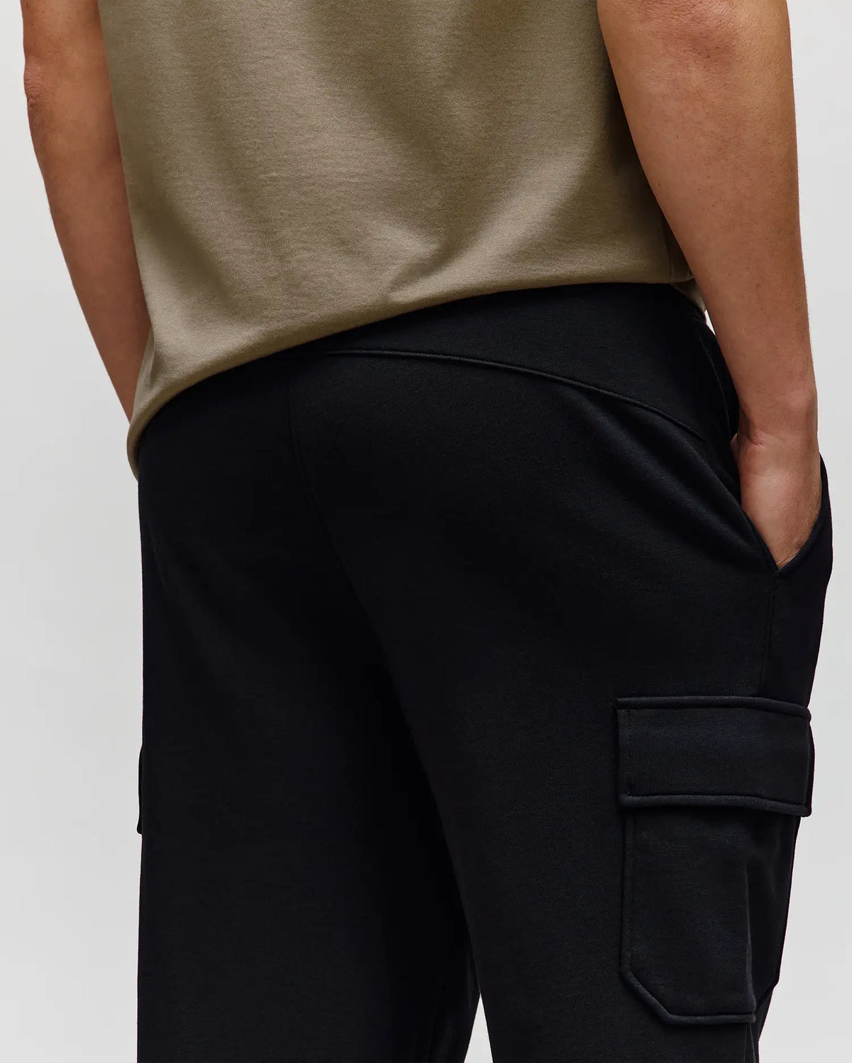 A person in Psycho Bunny’s MENS NASH CARGO SWEATPANT (MB0800010)—black cargo sweatpants with side pockets—stands with one hand in their pocket, waist-level from the back, blending comfort and practical style.