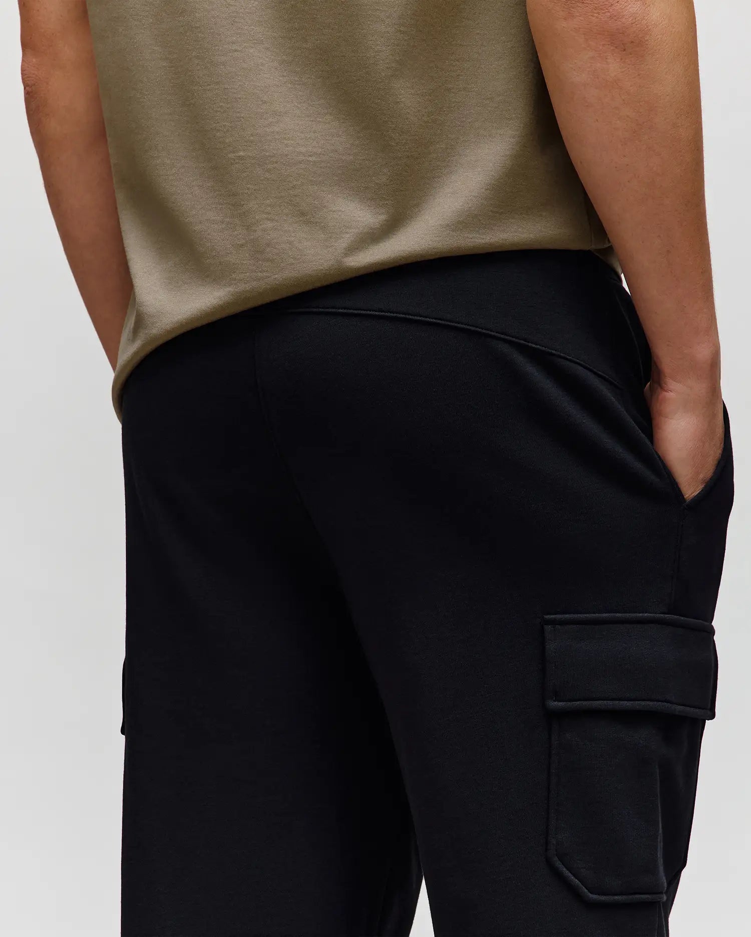 A person in Psycho Bunny’s MENS NASH CARGO SWEATPANT (MB0800010)—black cargo sweatpants with side pockets—stands with one hand in their pocket, waist-level from the back, blending comfort and practical style.
