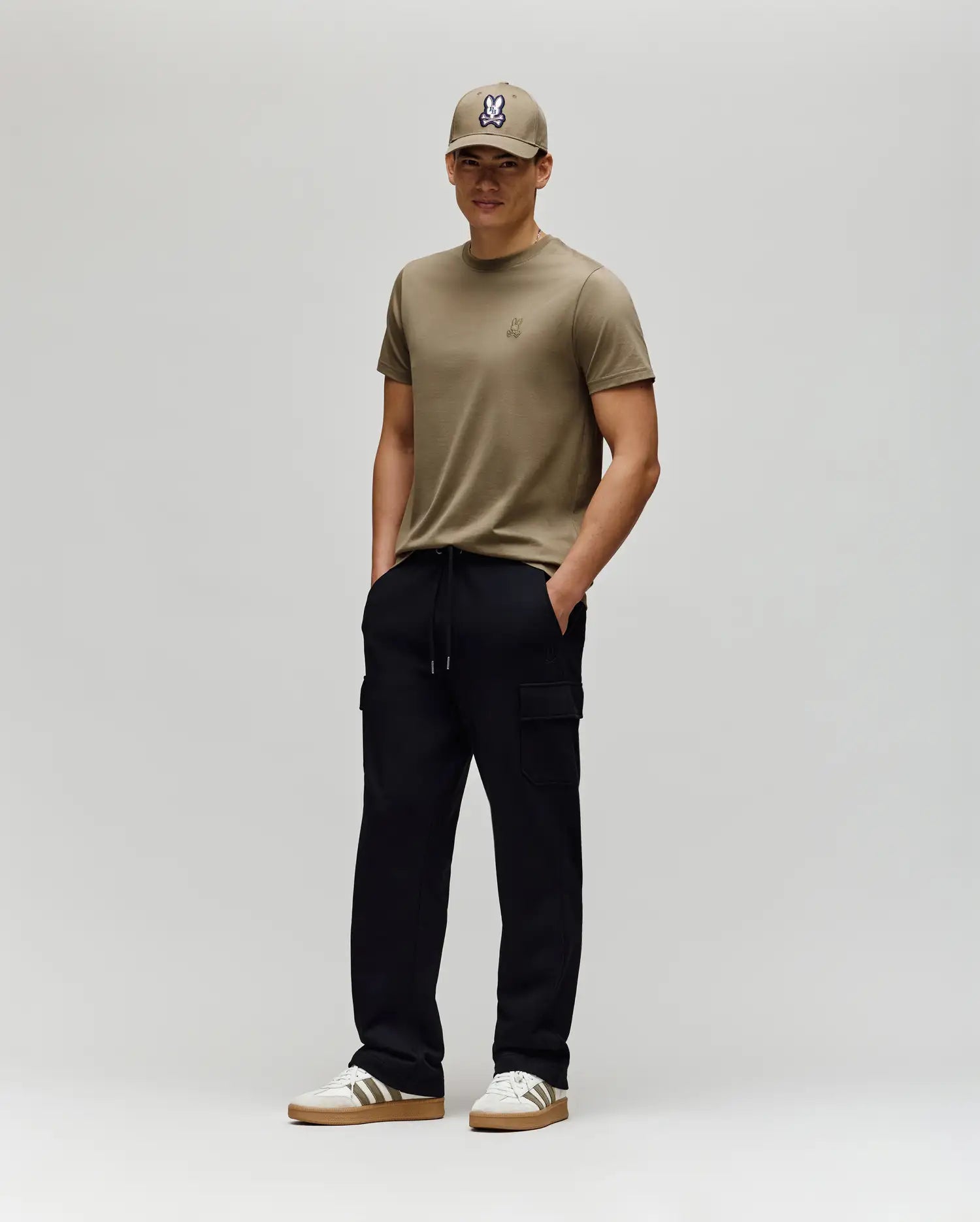 A man stands against a plain background wearing a tan cap, matching t-shirt, Psycho Bunny MENS NASH CARGO SWEATPANT - MB0800010 with cargo pockets, and white sneakers with brown soles. His hands are in his pockets and he smiles slightly.