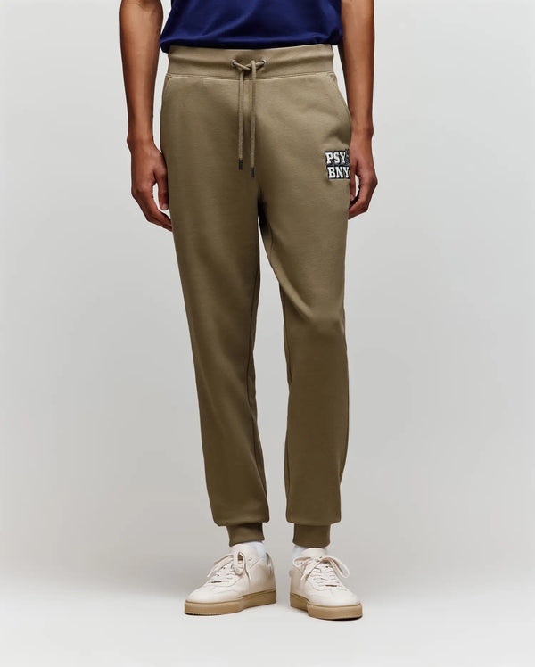 A person wearing Psycho Bunny MENS WADE SWEATPANT - MB0800014 in olive green, with "PSY BNK" text on the left thigh, pairs them with a navy shirt and white sneakers, standing against a plain light gray background.