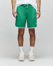 A person wearing Psycho Bunny MENS NICO MESH SHORT - MB0900004 in green with white trim, a white T-shirt, white crew socks, and white sneakers stands against a light gray background.