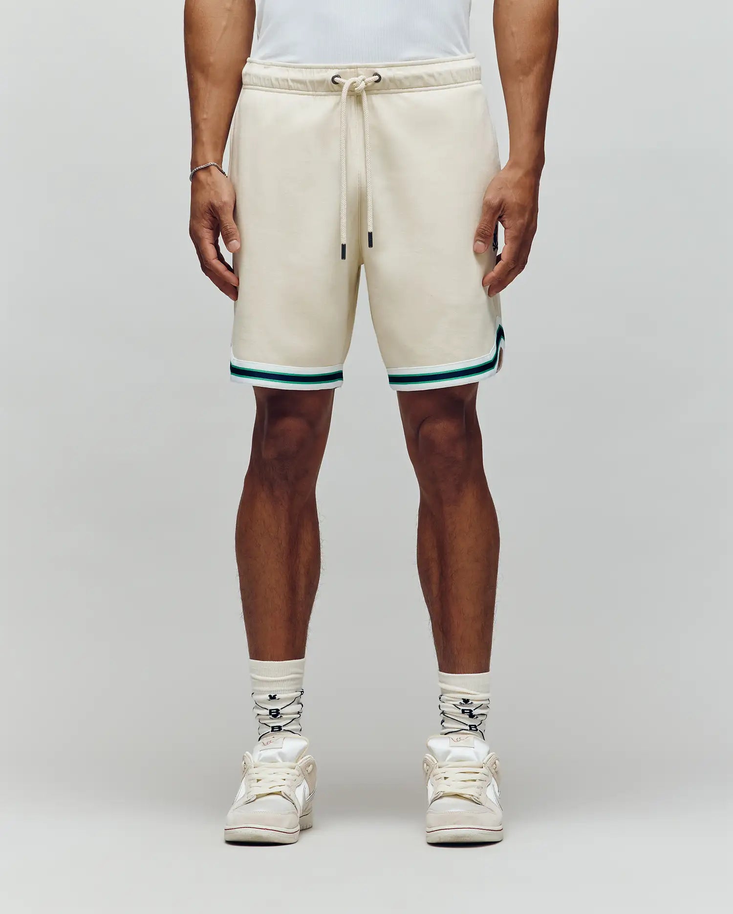 A person wearing Psycho Bunny’s MENS BASIL TAPED SHORT - MB0900005 in cream with teal and black trim, paired with white socks, white sneakers, and a white shirt stands against a plain light gray background.
