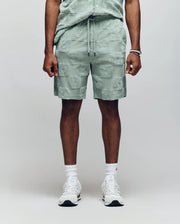 A person (head not visible) stands against a plain background wearing Psycho Bunny MENS KELVIN TOWEL TERRY SWEATSHORT - MB0900006 in light green jacquard terry cloth with matching shirt, white socks, white sneakers, and a bracelet.