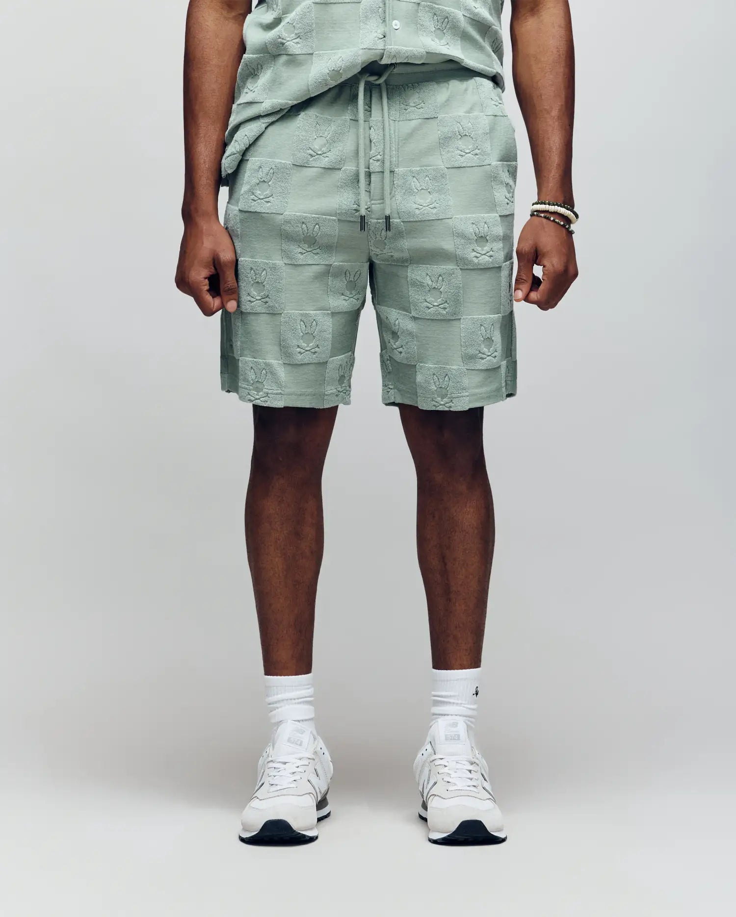 A person (head not visible) stands against a plain background wearing Psycho Bunny MENS KELVIN TOWEL TERRY SWEATSHORT - MB0900006 in light green jacquard terry cloth with matching shirt, white socks, white sneakers, and a bracelet.