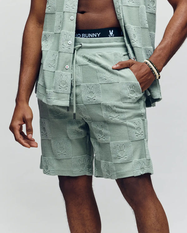 A person wears Psycho Bunny’s MENS KELVIN TOWEL TERRY SWEATSHORT (MB0900006) in light green jacquard terry cloth with an embroidered Checkered Bunny, black “Psycho Bunny” waistband, and matching top; image shows from shoulders to knees.