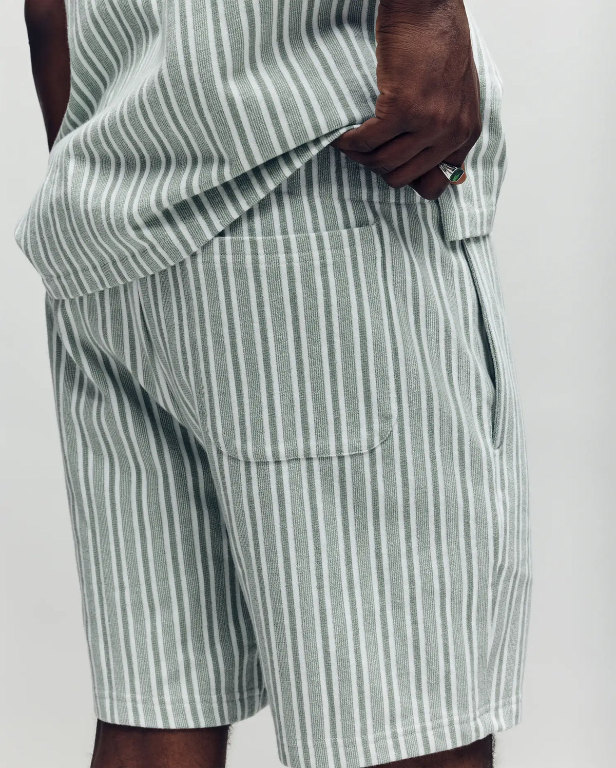 A close-up shows a person in Psycho Bunny's MENS ROBERT KNIT STRIPE SHORT - MB0900007, featuring light gray and white vertical stripes, a matching shirt, and an embroidered bunny near the back pocket. The background is plain and light-colored.