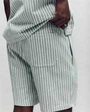 A close-up shows a person in Psycho Bunny's MENS ROBERT KNIT STRIPE SHORT - MB0900007, featuring light gray and white vertical stripes, a matching shirt, and an embroidered bunny near the back pocket. The background is plain and light-colored.
