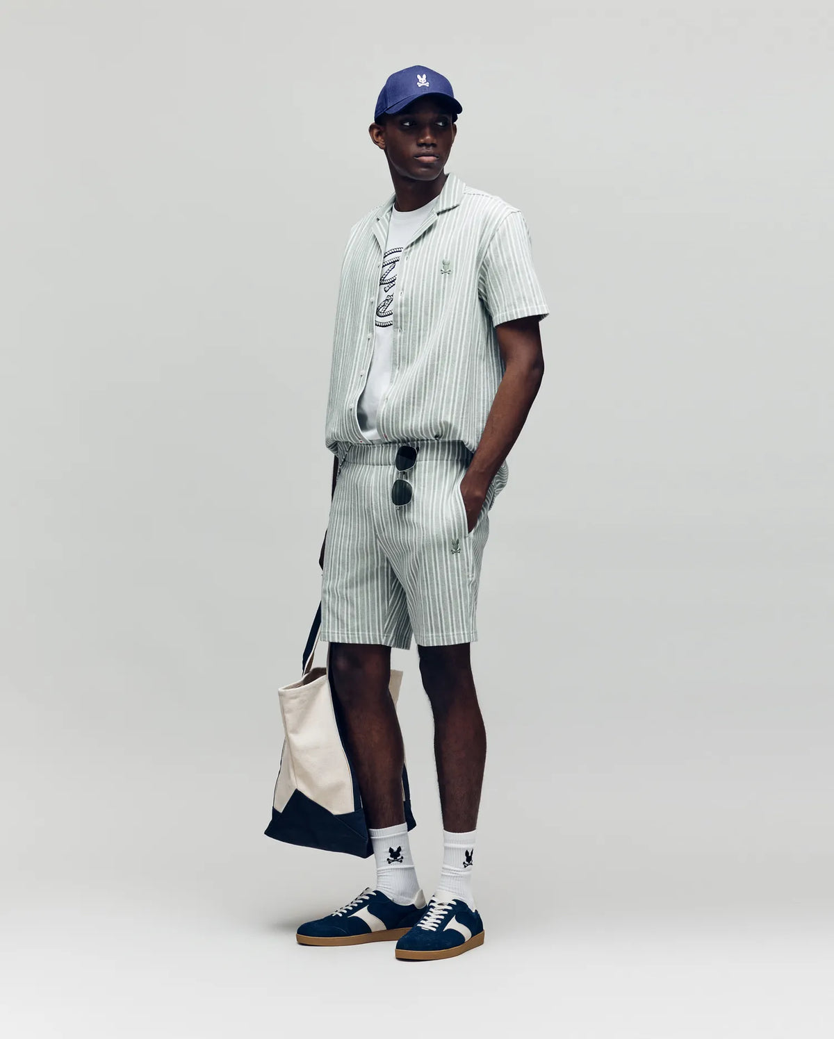 A man stands against a plain background wearing Psycho Bunny’s MENS ROBERT KNIT STRIPE SHORT (MB0900007) set, with a gray tee, navy and white sneakers, white socks, blue cap, sunglasses, and carrying a beige tote bag.