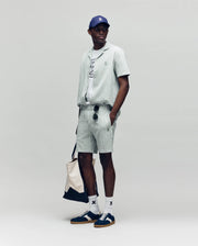 A man stands against a plain background wearing Psycho Bunny’s MENS ROBERT KNIT STRIPE SHORT (MB0900007) set, with a gray tee, navy and white sneakers, white socks, blue cap, sunglasses, and carrying a beige tote bag.