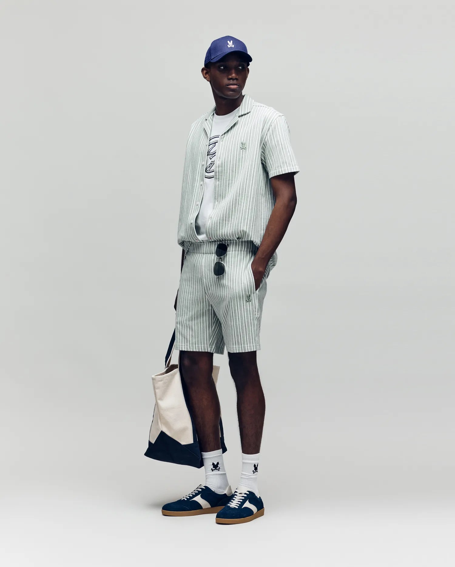 A man stands against a plain background wearing Psycho Bunny’s MENS ROBERT KNIT STRIPE SHORT (MB0900007) set, with a gray tee, navy and white sneakers, white socks, blue cap, sunglasses, and carrying a beige tote bag.