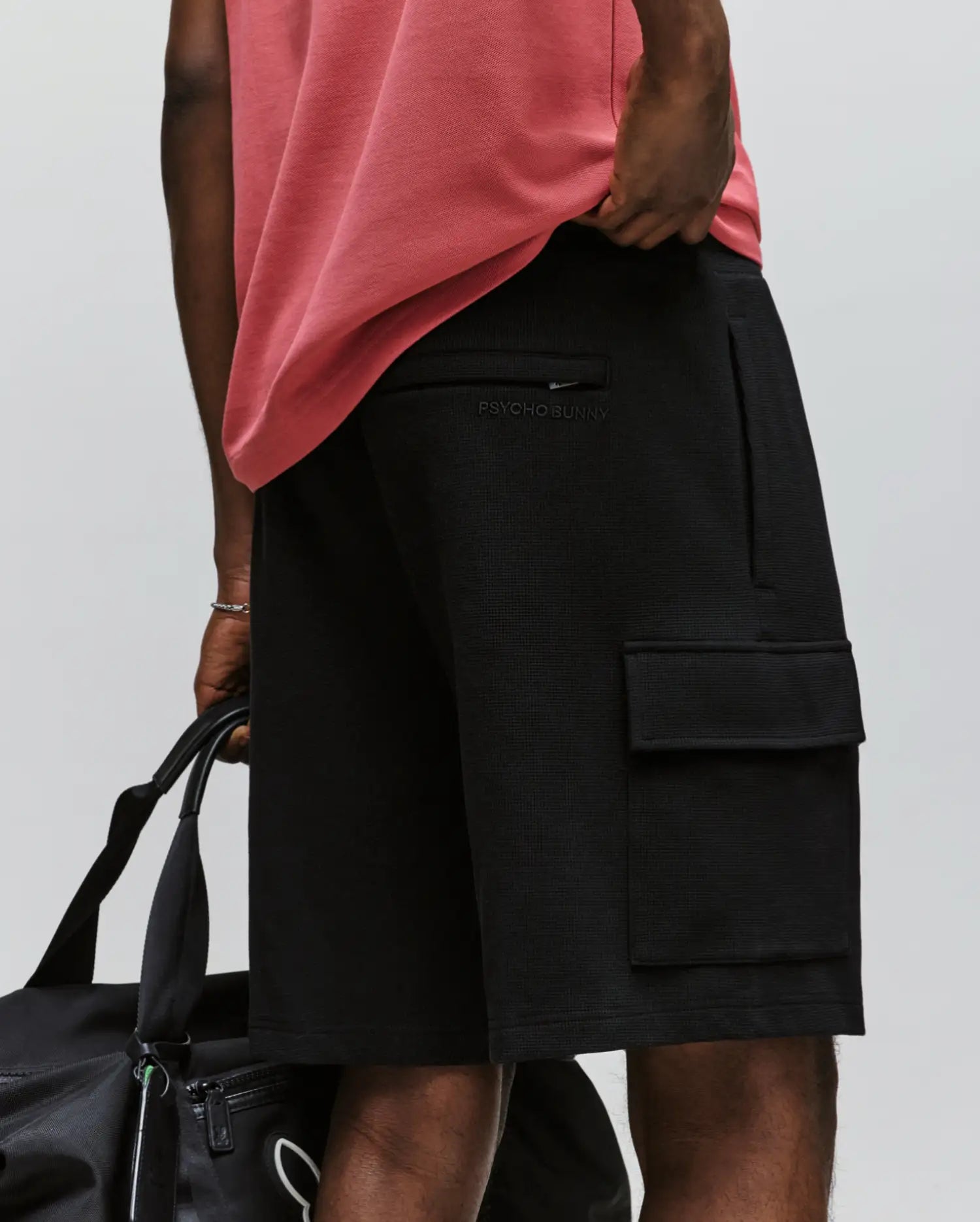 A person wears a pink shirt and Psycho Bunny MENS RON WAFFLE CARGO SHORT - MB0900012, black with zip/flap pockets and embroidered logo, holding a black duffel bag against a plain light background.