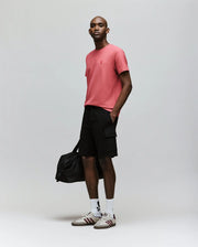 A man stands confidently in a pink tee, white socks, sneakers with embroidered logos, and Psycho Bunny MENS RON WAFFLE CARGO SHORT - MB0900012, holding a black duffel bag against a plain light gray background.