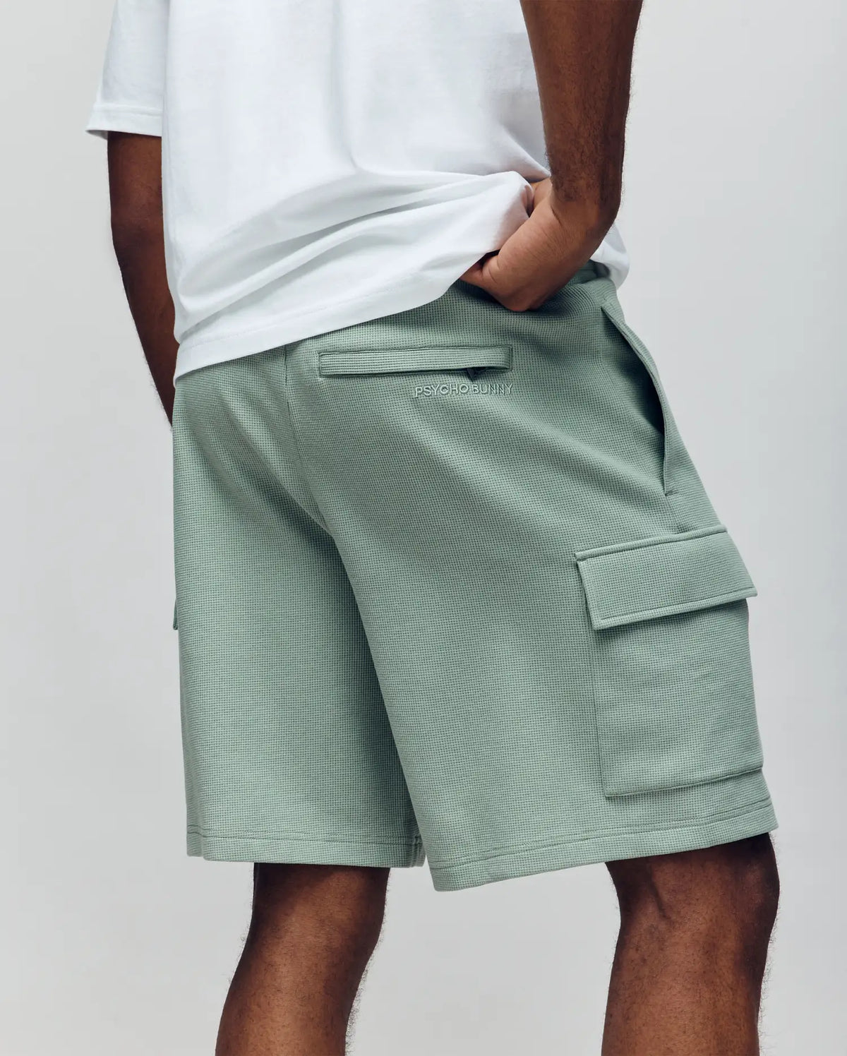 A person is seen from the back wearing a white waffle knit tee and Psycho Bunny MENS RON WAFFLE CARGO SHORT - MB0900012 in light green, featuring side and back pockets, embroidered logos, relaxed fit, and one hand in a pocket.