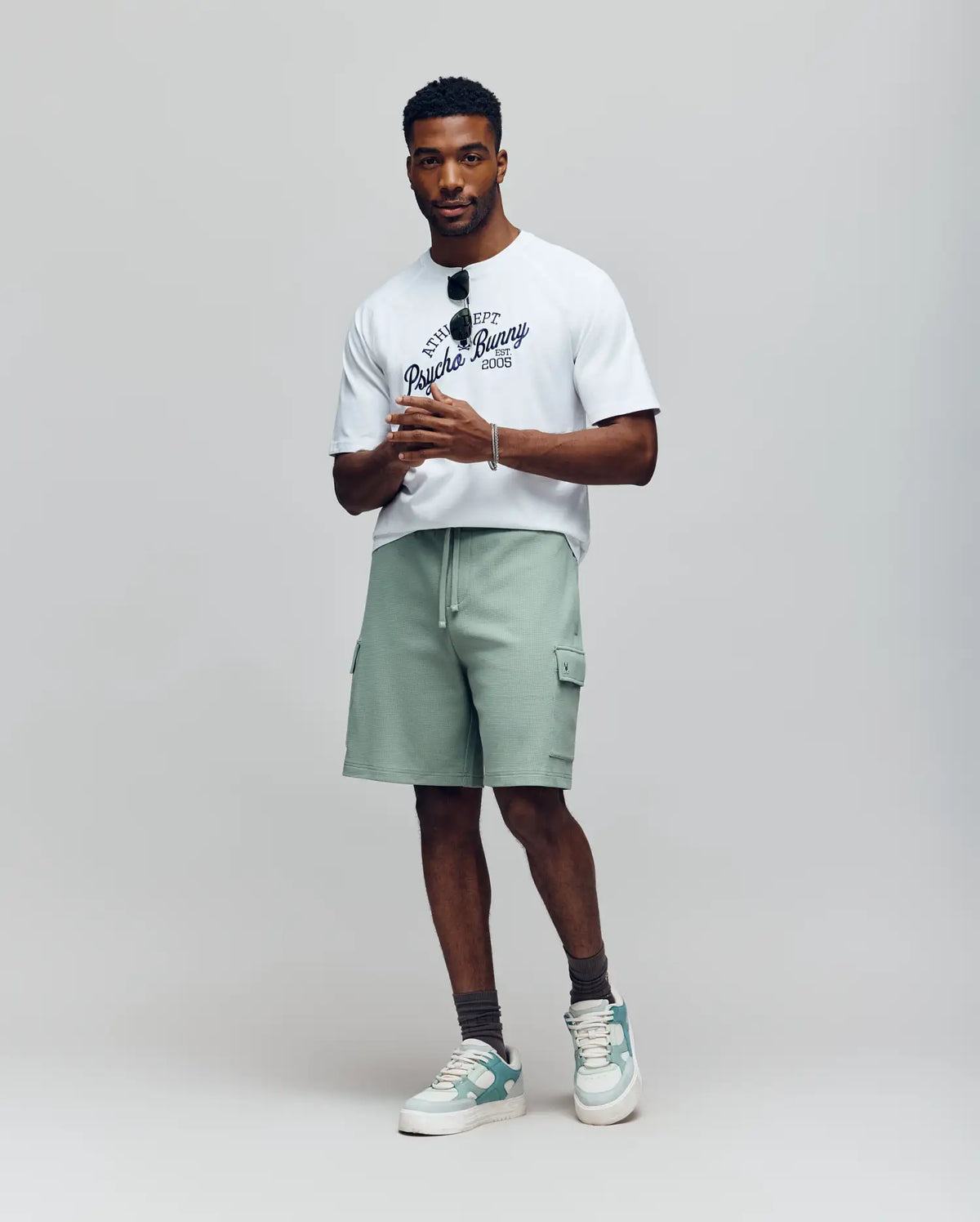A man stands against a plain background wearing a white graphic tee, gray socks, green and white sneakers, and Psycho Bunny’s MENS RON WAFFLE CARGO SHORT (MB0900012) in light green with embroidered logos. He holds sunglasses confidently.
