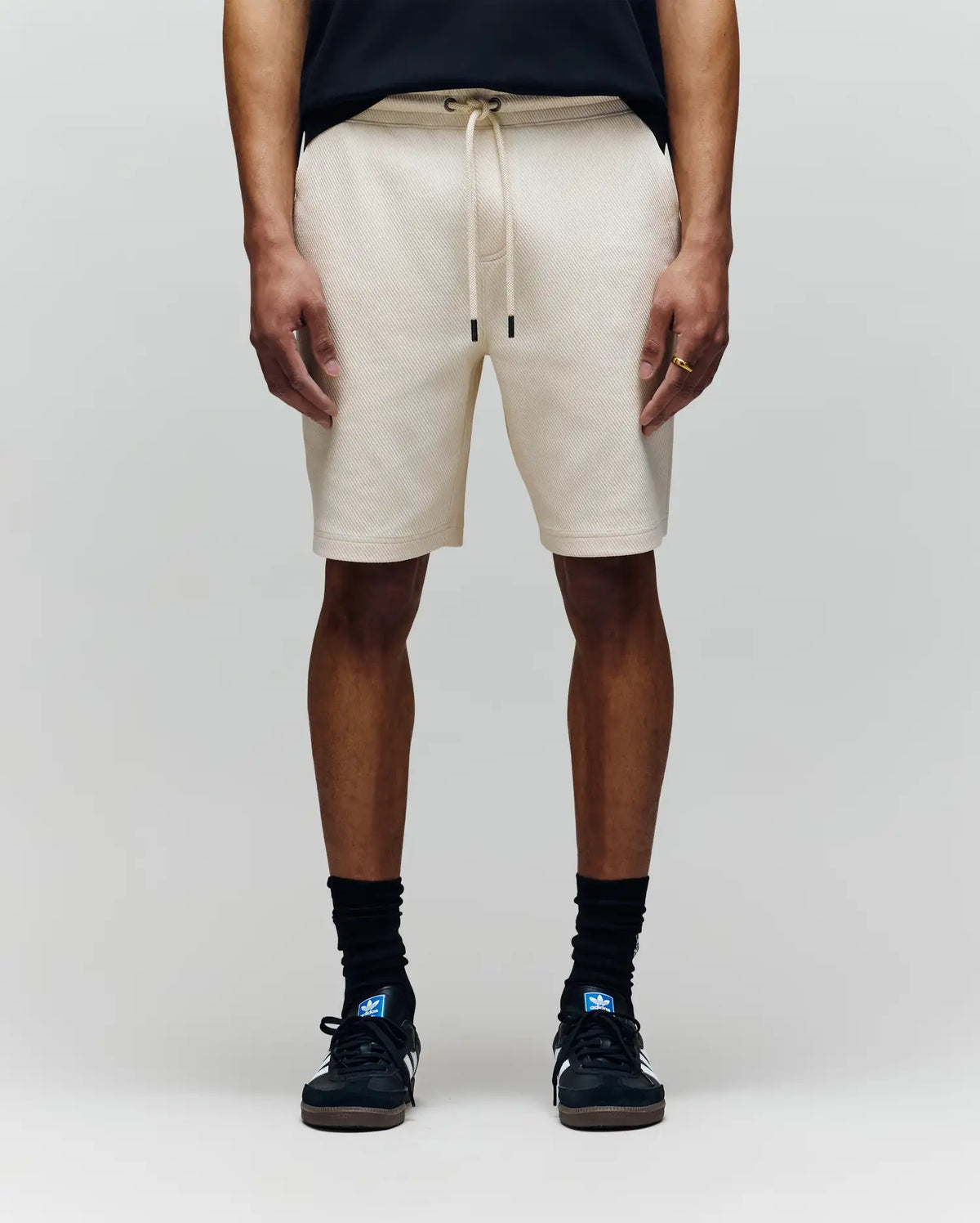 A person wearing Psycho Bunny MENS KAI SWEATSHORT - MB0900015 in beige with an embroidered Bunny logo, a black T-shirt, black crew socks, and black sneakers stands against a plain light gray background.