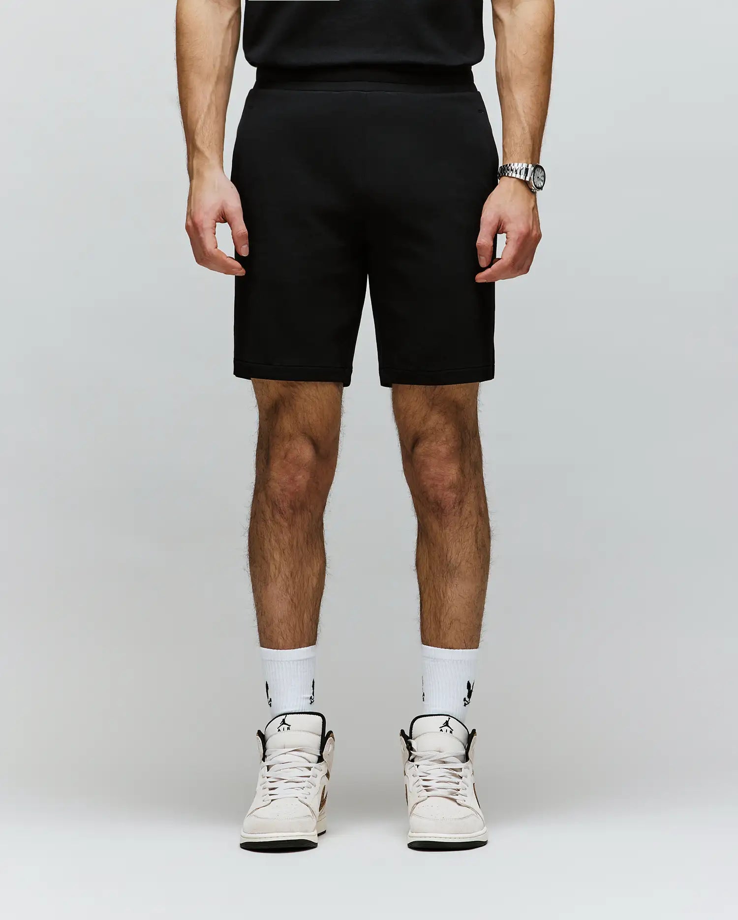 A person, shown from the shoulders down, wears Psycho Bunny MENS LAZSLO SWEATSHORT - MB0900016 in a stretch modal-cotton blend, paired with a black shirt, wristwatch, white logo socks, and white sneakers for an effortless casual look.