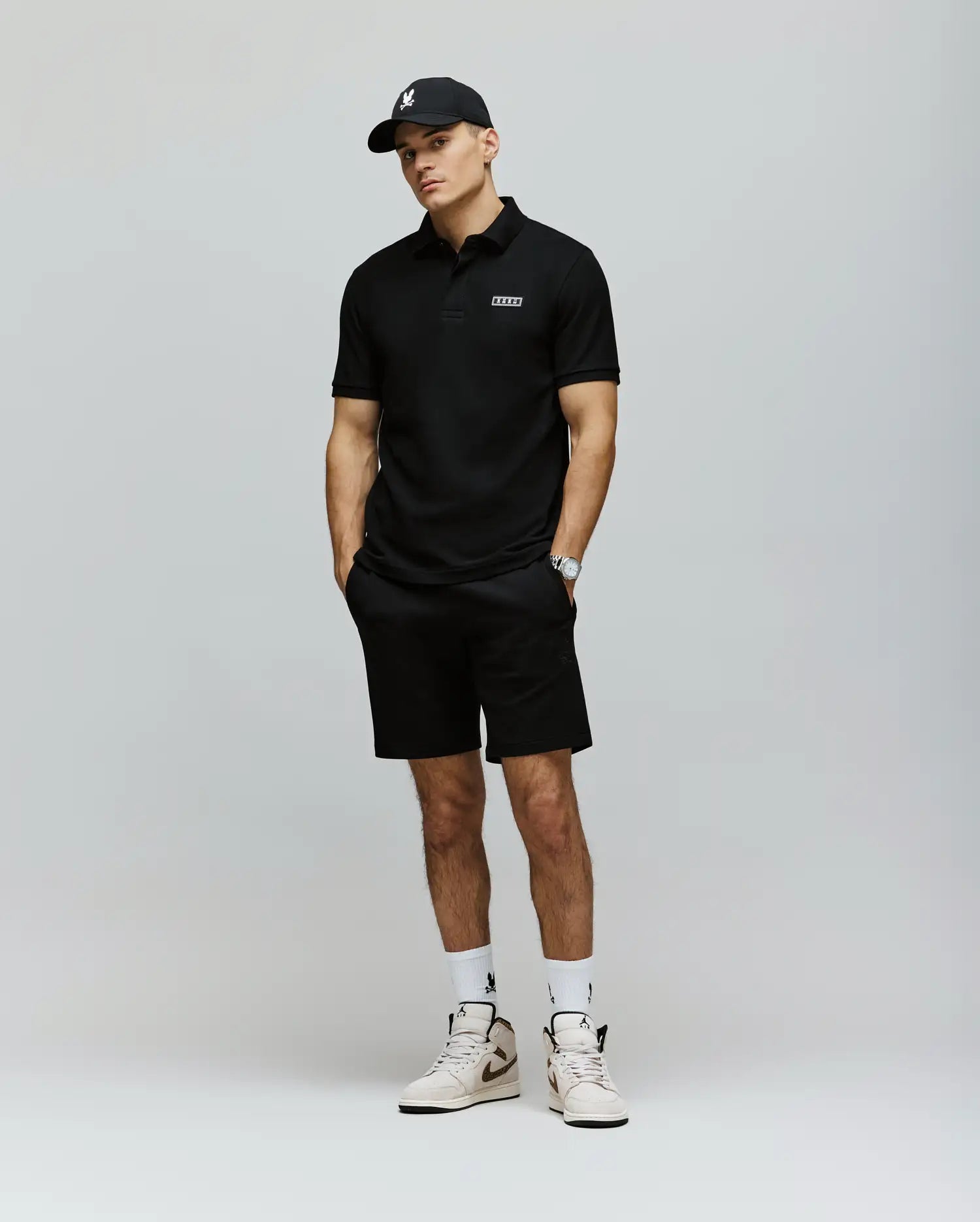 A man in a black polo, Psycho Bunny MENS LAZSLO SWEATSHORT - MB0900016, white socks and sneakers, and a black cap stands hands-in-pockets against a light gray background for a perfected casual look.