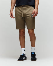 A person wearing Psycho Bunny MENS WADE SWEATSHORT - MB0900028 in tan, featuring an embroidered “FEVR” wordmark, paired with white socks and black Adidas sneakers, stands against a plain light background.