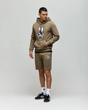 A man in Psycho Bunny MENS WADE SWEATSHORT - MB0900028 poses against a plain background, wearing an olive green hoodie, olive shorts with white text, white socks, and black Adidas sneakers for a classic athletic look.