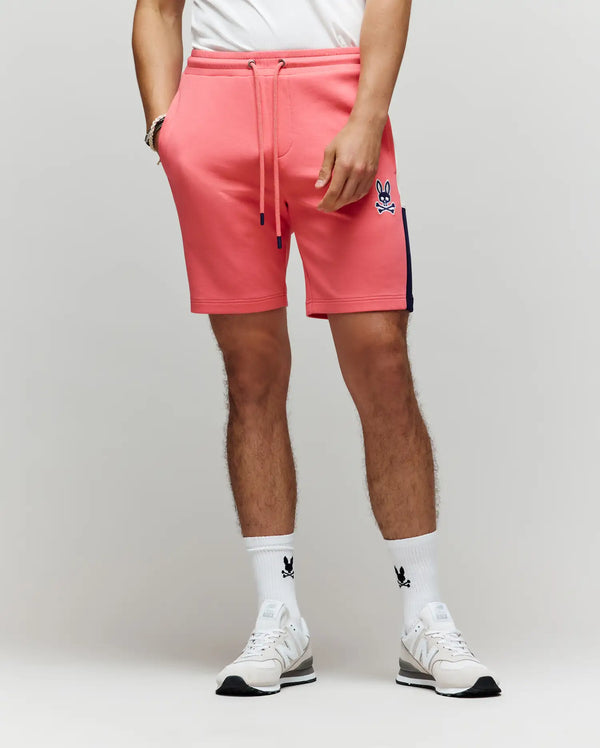 A person in Psycho Bunny's MENS DAWSON TRACK SHORT - MB0900032 in coral pink with a navy side stripe, plus a white tee and white bunny logo socks, showcases effortless street style and loungewear comfort against a plain background.