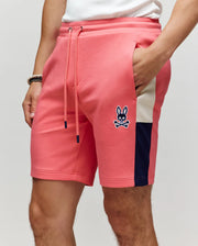 A person wears Psycho Bunny’s MEN'S DAWSON TRACK SHORT - MB0900032 in coral pink with navy and white side stripes, a drawstring waist, and bunny skull logo, styled with a white shirt, bracelets, and one hand in their pocket.