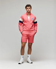 A man stands against a plain background wearing Psycho Bunny’s pink track jacket with navy and white sleeve stripes, matching MENS DAWSON TRACK SHORT (MB0900032), white socks, and sneakers, all accented by the signature bunny logo.