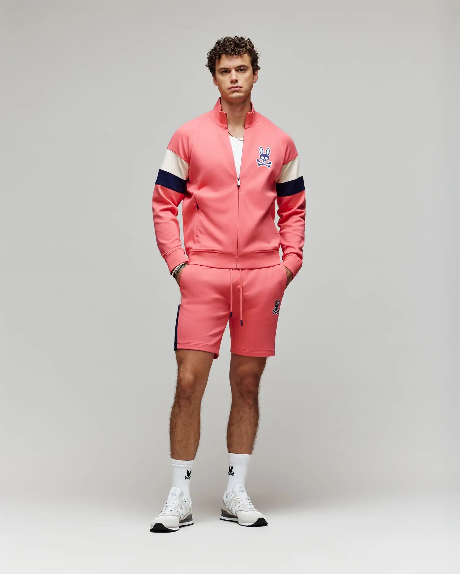 A man stands against a plain background wearing Psycho Bunny’s pink track jacket with navy and white sleeve stripes, matching MENS DAWSON TRACK SHORT (MB0900032), white socks, and sneakers, all accented by the signature bunny logo.