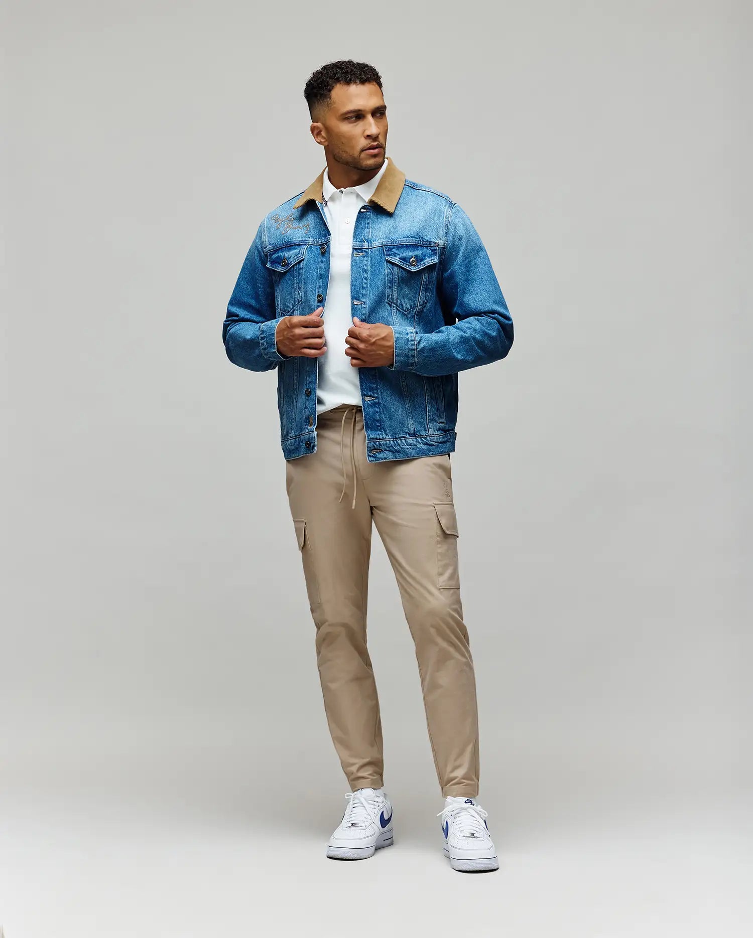 A man stands against a plain background wearing the Psycho Bunny MENS WADE DENIM TRUCKER JACKET - MO1100015 with a tan corduroy collar, paired with a white shirt, beige cargo pants, and white sneakers. He looks to the side, holding his jacket’s front.
