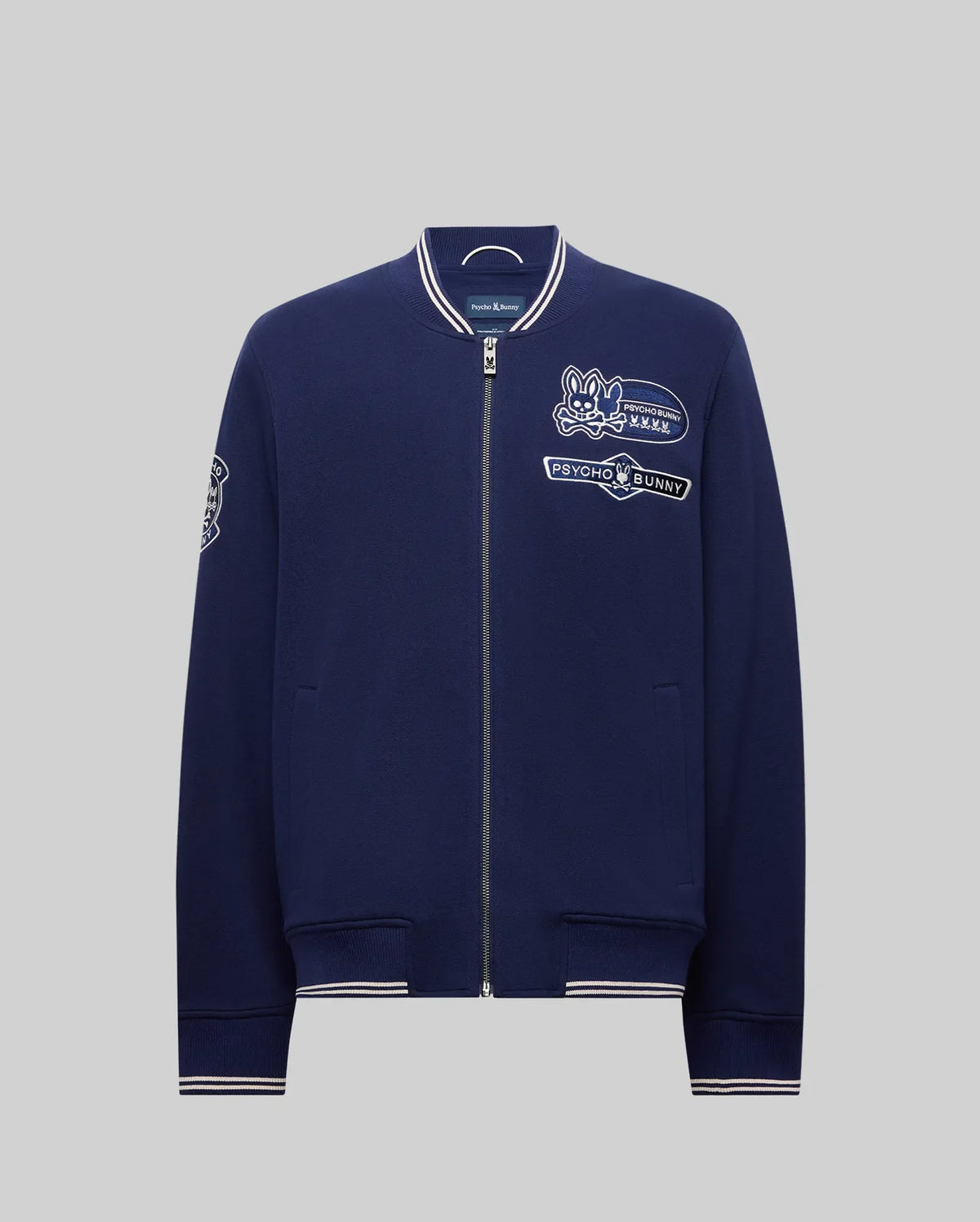 The Psycho Bunny MENS DESMOND PIQUE KNIT BOMBER - MO1100018 is a navy bomber with white trim, varsity-style details, and embroidered patches including a bunny logo, "Psycho Bunny" text on the chest, and a sleeve patch.