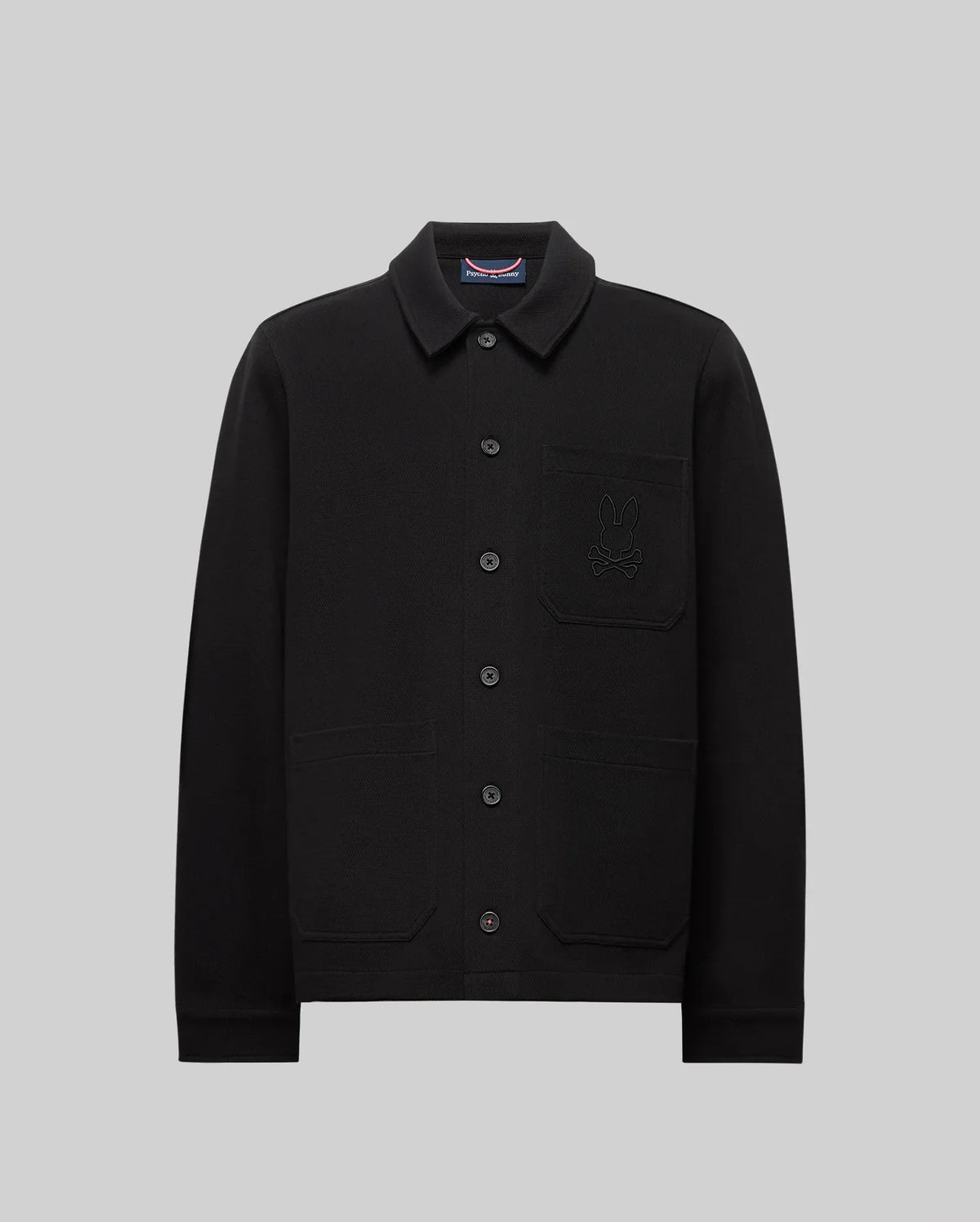 The Psycho Bunny MENS AVEN KNIT SHACKET (MO1100035) is a black button-up knit shacket with a collar, three front pockets, and an embroidered cartoon bunny face on the left chest pocket, shown on a plain gray background.