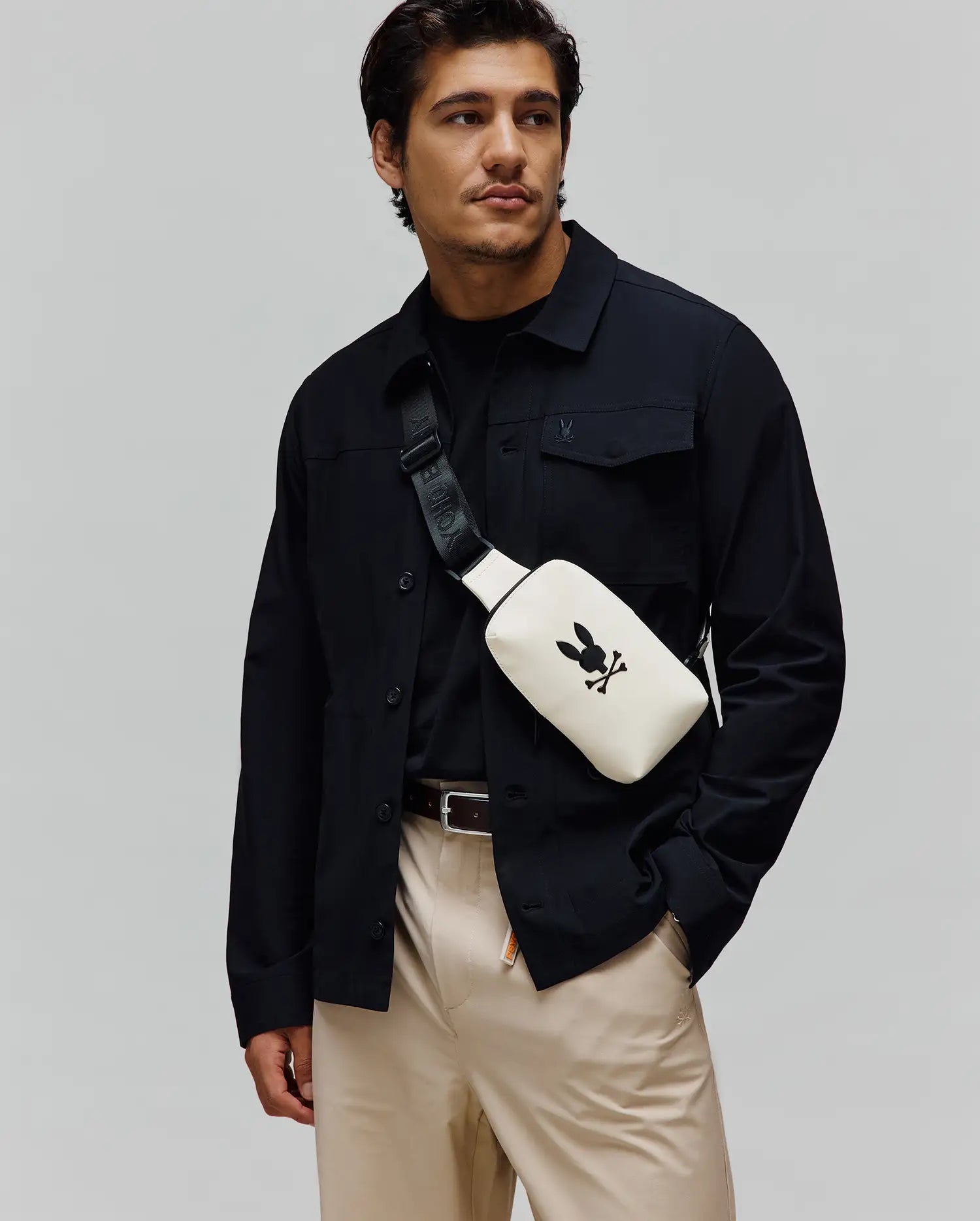 Mens Rubberized Crossbody in cream with a large black Psycho Bunny skull logo, worn with a black collared shirt jacket featuring a small embroidered bunny, a black crew neck tee, and khaki pants.