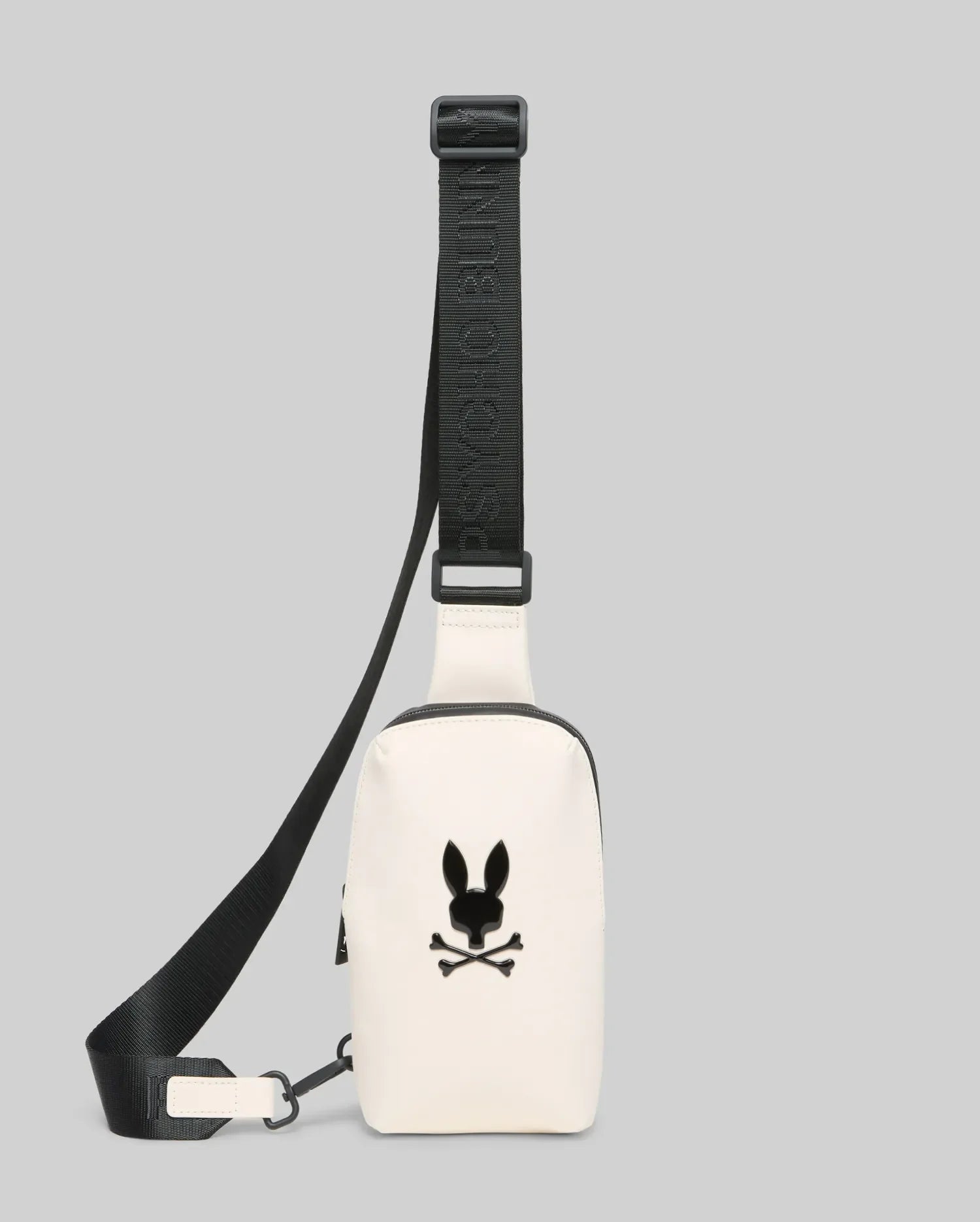Mens Rubberized Crossbody in cream, featuring a smooth finish, a prominent black Psycho Bunny skull logo on the front, and an adjustable black strap with repeated brand text.