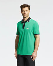 A man wearing the Psycho Bunny MENS CLAYTON PIQUE POLO (MT0100004), designed in a breathable Pima cotton, stands against a plain background. The green shirt is colorblocked with white and black on the collar and sleeves, featuring a black collar and logo on the chest.