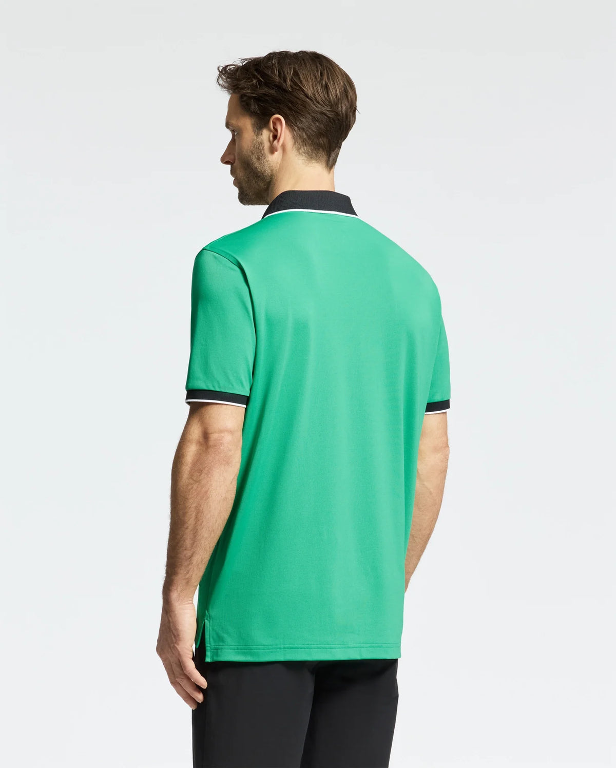 A man with brown hair stands with his back to the camera wearing a Psycho Bunny MENS CLAYTON PIQUE POLO - MT0100004. This green polo, featuring a black collar and sleeve trim, is made of premium Pima cotton for a breathable fit. He pairs it with black pants against a light gray background.