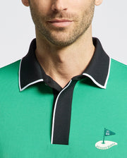Wearing the MEN'S CLAYTON PIQUE POLO by Psycho Bunny, this person sports a premium Pima cotton green polo with a black collar and white trim. It features an embroidered flag-and-rabbit logo with "Psycho Bunny" text, while their beard and lower face are partially visible.
