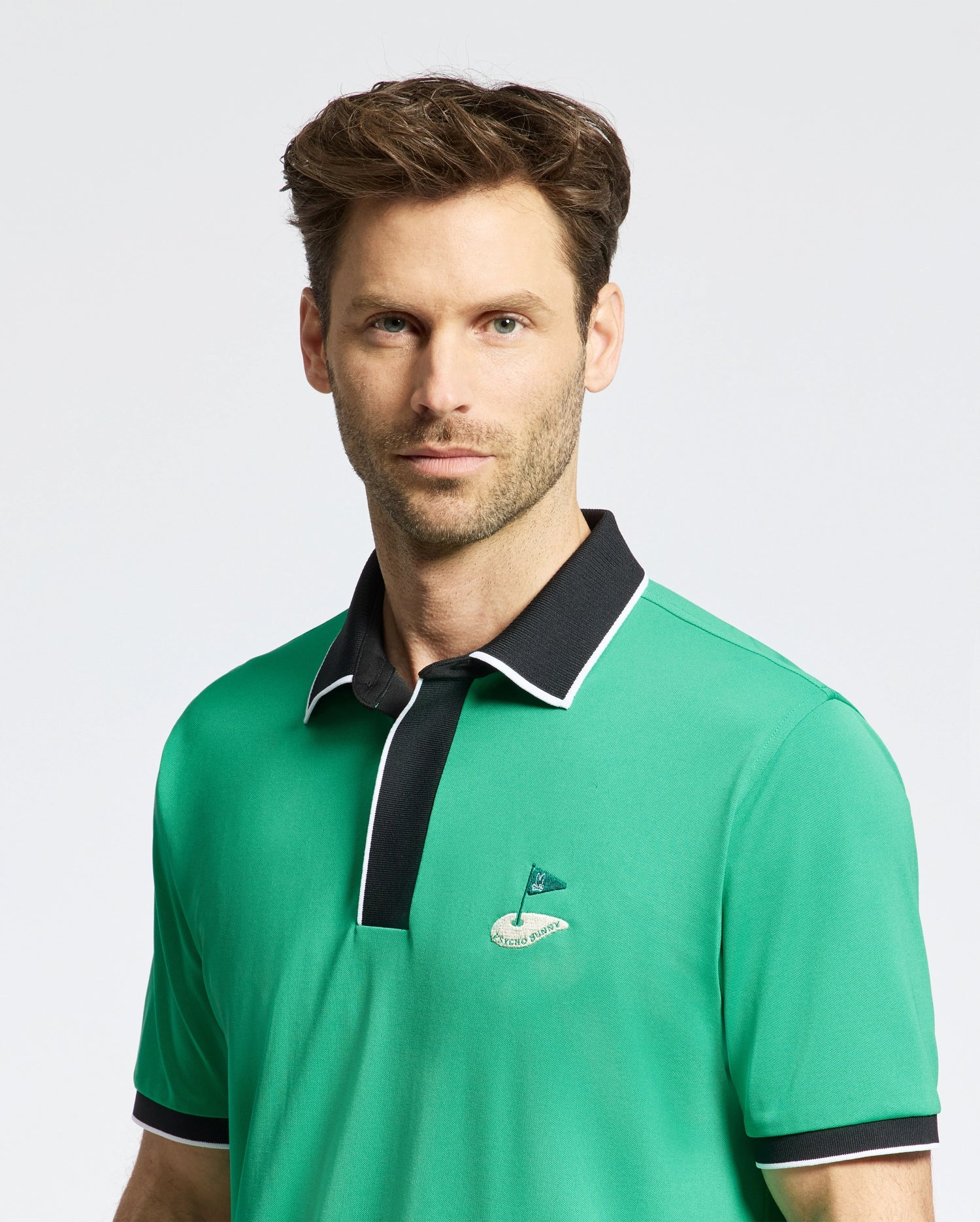 A person stands against a plain light background, wearing a Psycho Bunny MENS CLAYTON PIQUE POLO - MT0100004. This green polo features a black collar and small embroidered flag design, crafted from premium Pima cotton for breathable comfort.