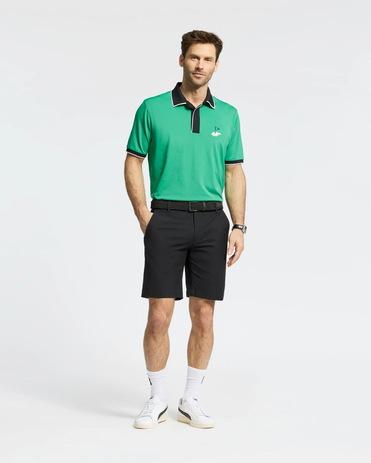 A man in a Psycho Bunny MENS CLAYTON PIQUE POLO - MT0100004, made of breathable Pima cotton with green and black colorblocked details and a small emblem, stands against a white background. He's wearing black shorts, white sneakers, and sports a watch on his left wrist with one hand in his pocket.